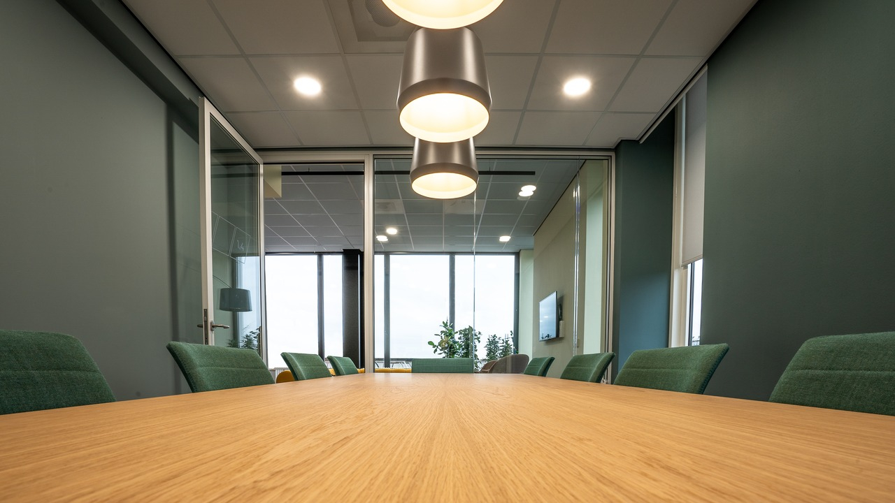 A brown table surrounded with green chairs under lamps in a room with glass acoustic partition wall